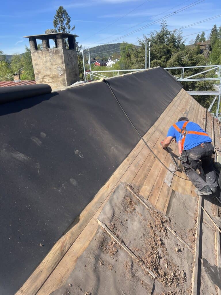 Taktekker jobber med å legge takbelegg på et skrånende tak med utsikt over landskapet. Faglig og erfarent håndverk for holdbare takløsninger.